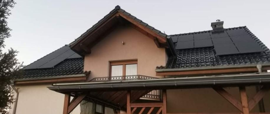 Panele na dachu dwukondygnacyjnego domu z drewnianą werandą, widoczne rynny i komin.