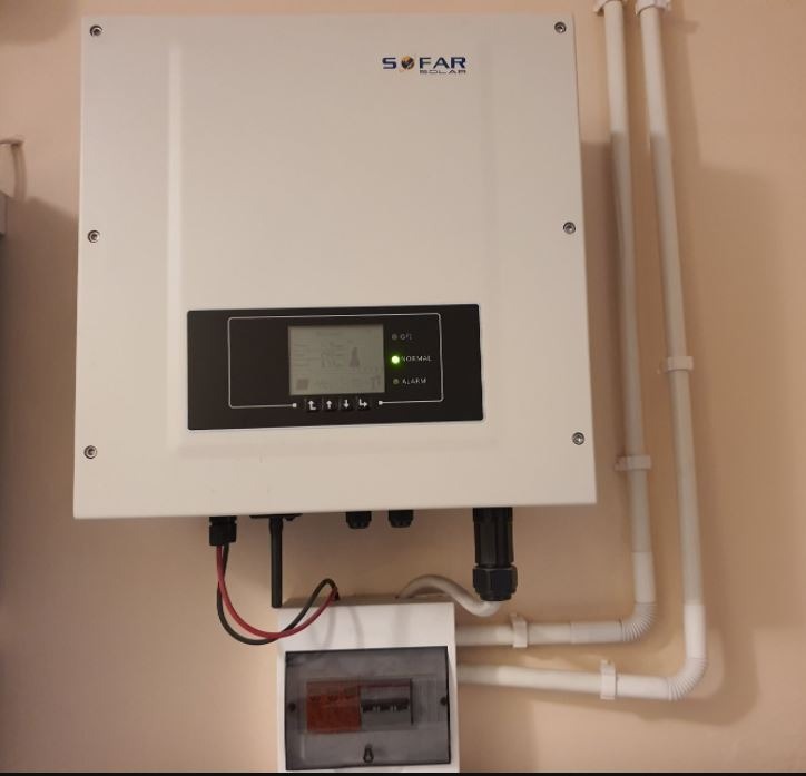 Biały falownik Sofar Solar zamontowany na beżowej ścianie, z widocznymi przewodami i skrzynką bezpiecznikową poniżej, oraz rurami instalacyjnymi po prawej stronie.