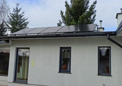 Czarny panel słoneczny na dachu jednopiętrowego domu z białym tynkiem i ciemnymi ramami okiennymi, widoczne drzewa w tle.