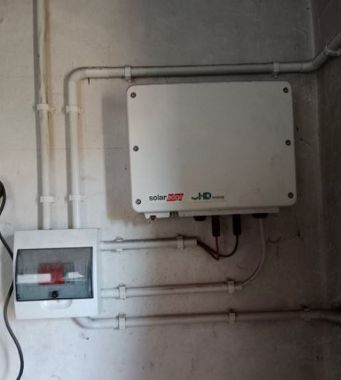 Biała skrzynka inwertera SolarEdge HD-Wave zamontowana na szarej ścianie obok skrzynki bezpiecznikowej i rur instalacyjnych.