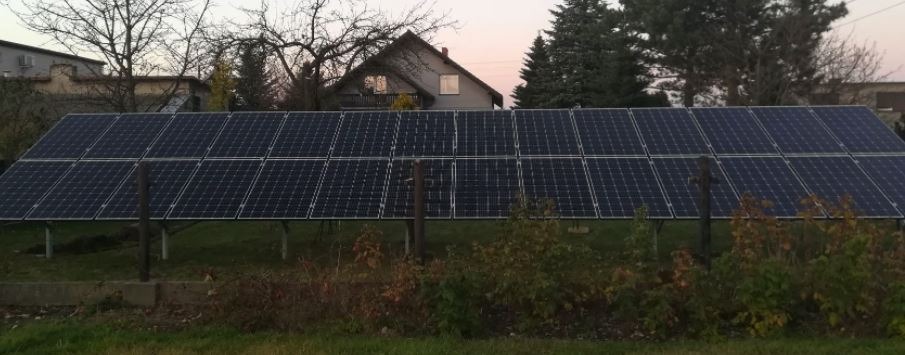 Instalacja paneli słonecznych na metalowej konstrukcji, widoczna zza niskiego ogrodzenia na tle domu i drzew.