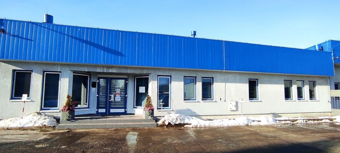 Budynek biurowy z niebieskim dachem blaszanym, widoczne okna, drzwi wejściowe, donice z kwiatami i resztki śniegu na parkingu.