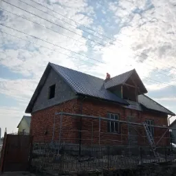 Ceglany dom w trakcie budowy z częściowo ukończonym, ciemnym dachem z blachy falistej; widoczny dekarz pracujący na dachu, rusztowania przy ścianach i niezabudowane poddasze.