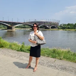 Kobieta w okularach przeciwsłonecznych, białej bluzce i czarnej spódnicy pozuje z torebką w kropki na tle Wisły z widokiem na most i Stadion Narodowy w Warszawie.