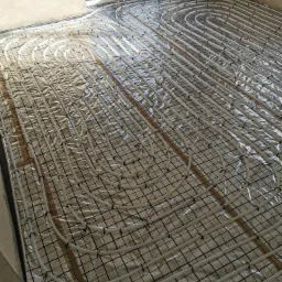 Białe rury ogrzewania podłogowego ułożone w pętle na srebrnej folii izolacyjnej, przymocowane do czarnej siatki na podłożu w surowym wnętrzu budynku.