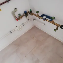 Kompleksowy węzeł wodomierzowy z licznymi zaworami, filtrem i rurami z tworzywa sztucznego, zamontowany na ścianie w pomieszczeniu z beżowymi płytkami podłogowymi.