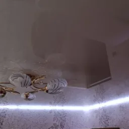 Błyszczący, kremowy sufit napinany z widocznym odbiciem wzoru tapety i ozdobnym, złotym żyrandolem z białymi kloszami w paski, oświetlony listwą LED przy ścianie z tapetą w kwiatowe wzory.