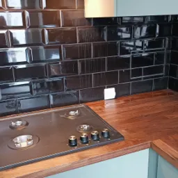 Czarne, błyszczące płytki typu metro na ścianie nad drewnianym blatem kuchennym w Sosnowcu, z zamontowaną płytą gazową z czarnymi pokrętłami.