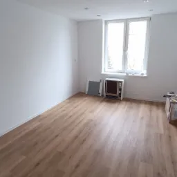 Jasne wnętrze po remoncie z panelami podłogowymi w odcieniach dębu, białymi ścianami i sufitami z wbudowanym oświetleniem punktowym. Przy oknie stoi grzejnik i płyta zabezpieczająca.