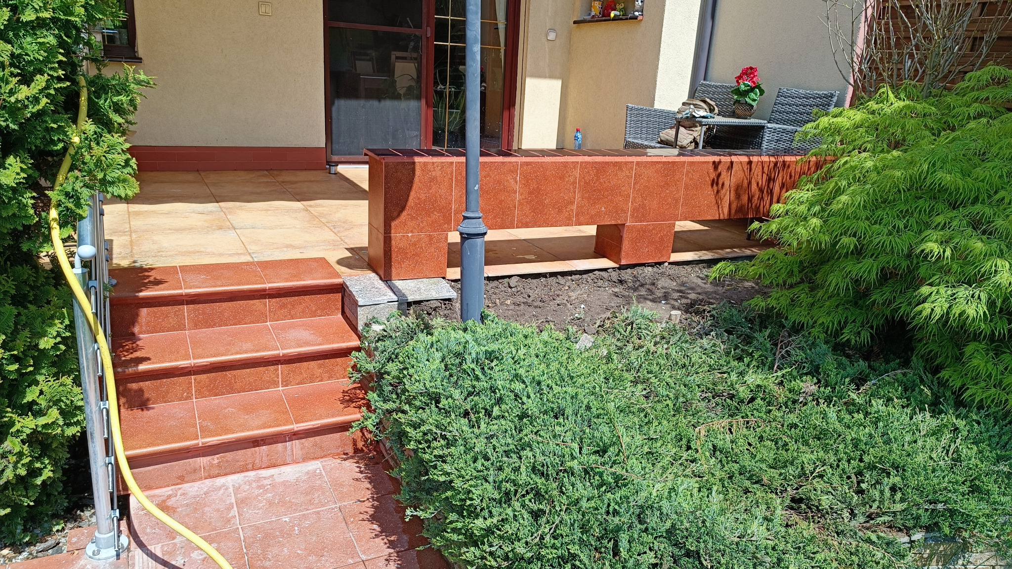 Wykończony taras z czerwonej terakoty z trzema schodami, balustradą z żółtym wężem ogrodowym, oraz niskim murem z terakoty, za którym widać meble ogrodowe i zieleń.