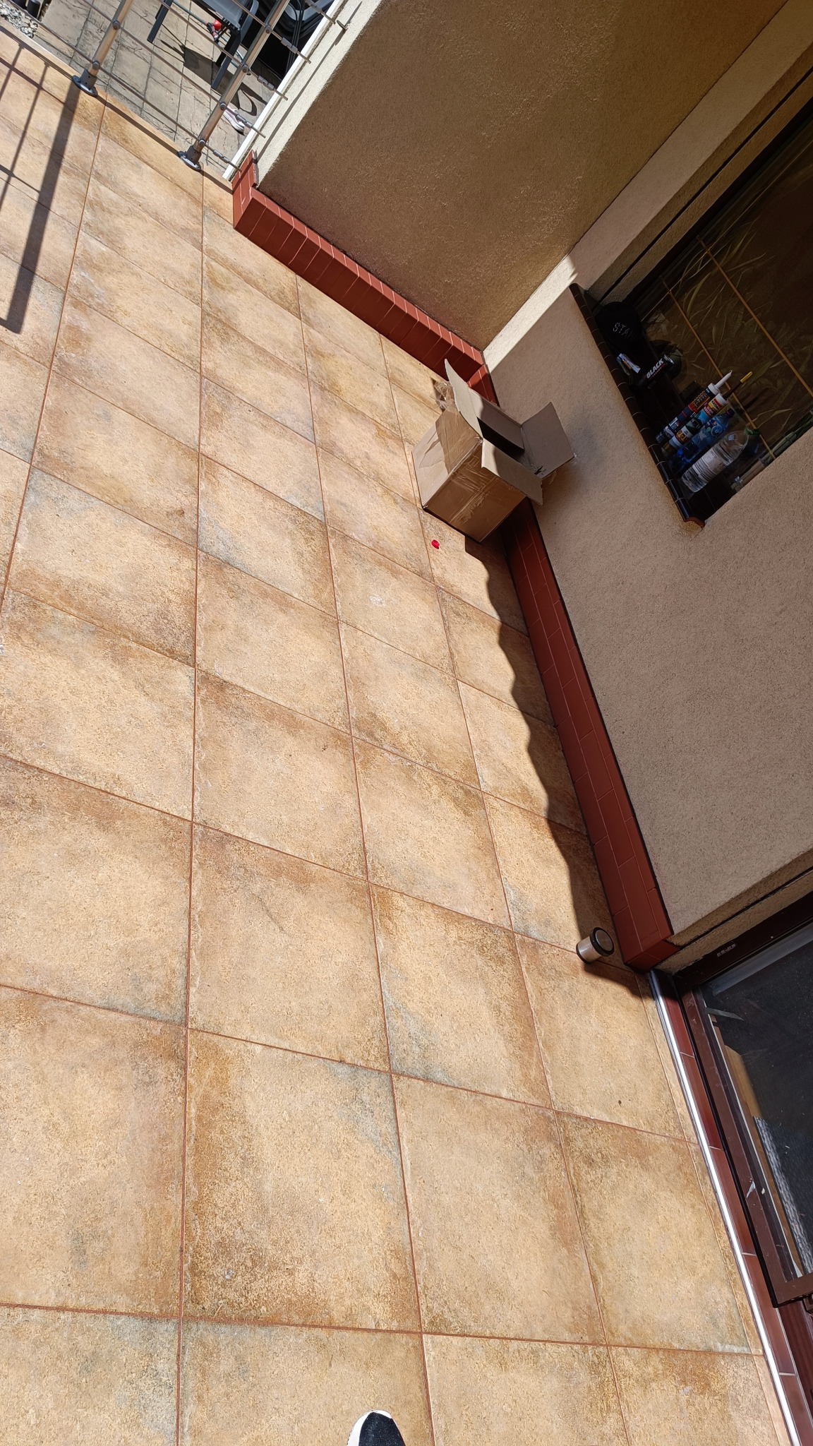 Beżowe płytki podłogowe na balkonie z otwartym kartonem i akcesoriami do glazury w tle, widok z góry.