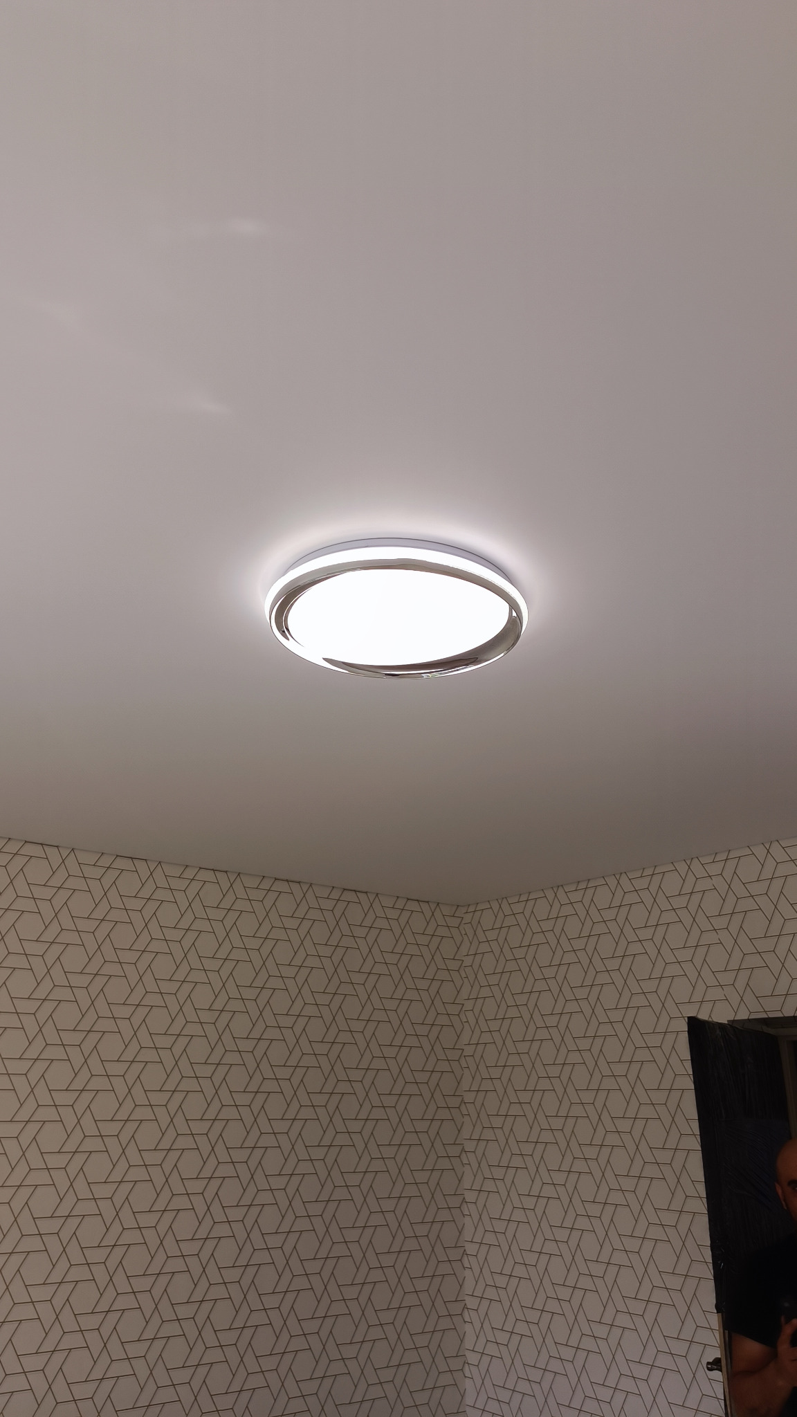 Okrągła lampa sufitowa LED z chromowanym wykończeniem, zamontowana na białym suficie w pomieszczeniu z tapetą w geometryczne wzory.