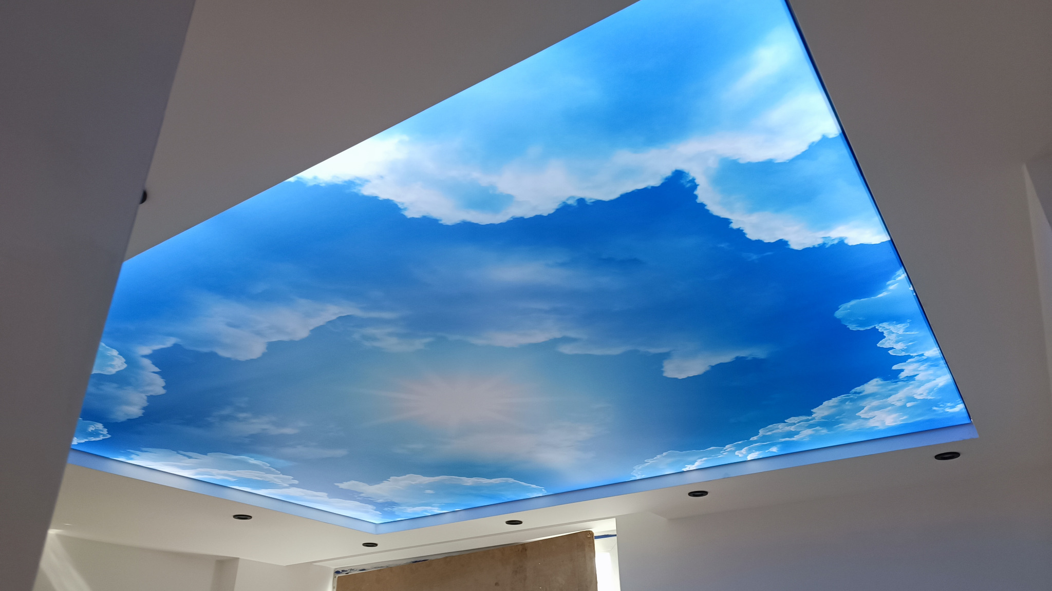 Niebieski sufit napinany z grafiką chmur i słońca, oświetlony od tyłu, w nowoczesnym wnętrzu.