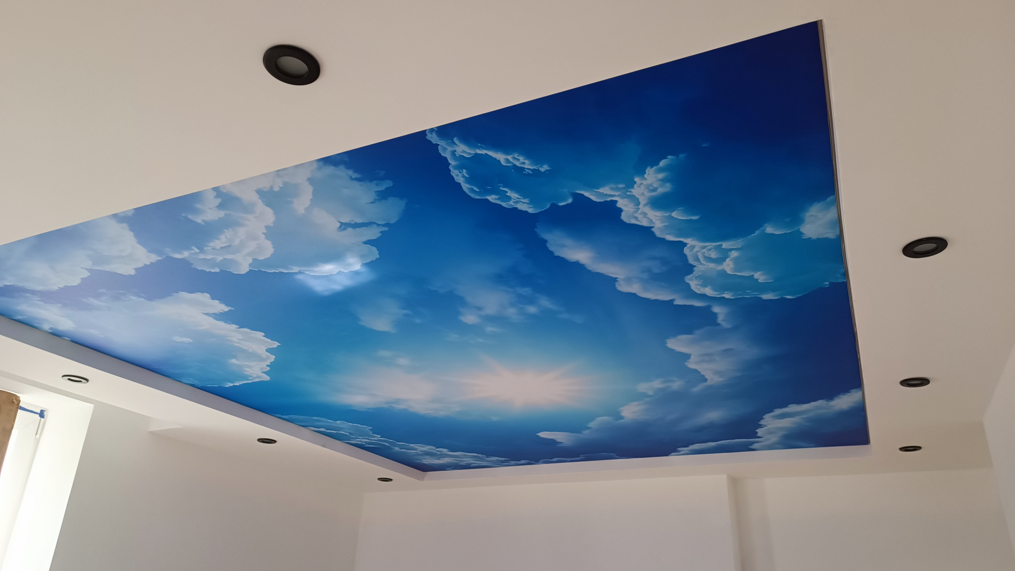 Błękitne niebo z chmurami i promieniami słońca na suficie napinanym, oświetlenie punktowe w białym pomieszczeniu.