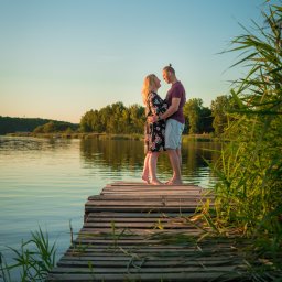 Mateusz Pudło - Para na drewnianym pomoście nad jeziorem o zachodzie słońca, kobieta w kwiecistej sukience obejmowana przez mężczyznę, widok na spokojną wodę i trzciny.