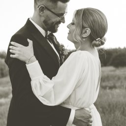 Mateusz Pudło - Czarno-białe zdjęcie pary młodej, obejmującej się i patrzącej sobie w oczy na tle łąki, panna młoda w sukni z bufiastymi rękawami, pan młody w garniturze i okularach.