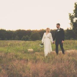 Mateusz Pudło - Para młoda trzymająca się za ręce spaceruje po łące w promieniach zachodzącego słońca, panna młoda w zwiewnej sukni z bukietem, pan młody w klasycznym smokingu z muszką.