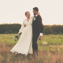 Mateusz Pudło - Para młoda w objęciach na łące o zachodzie słońca; panna młoda w zwiewnej sukni z bukietem białych róż, pan młody w klasycznym smokingu z muszką i butonierką.