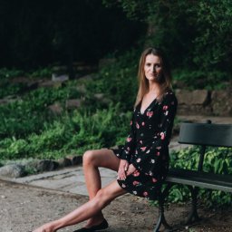 Mateusz Pudło - Kobieta w kwiecistej sukience siedzi na ławce w parku, z nogami skrzyżowanymi w kostkach i dłońmi splecionymi na kolanie.