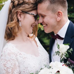 Mateusz Pudło - Uśmiechnięta para młoda, panna młoda w okularach, z warkoczem i welonem, pan młody w bordowej muszce, zbliżeni twarzami, bukiet z białych róż.