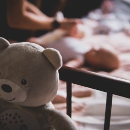 Mateusz Pudło - Pluszowy miś z aplikacją gwiazdek w łóżeczku, w tle osoba przewija niemowlę na różowej pościeli.