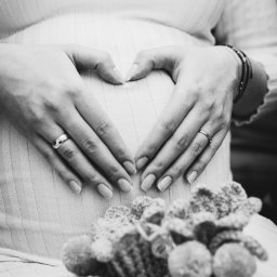 Mateusz Pudło - Czarno-białe zdjęcie kobiety w ciąży, dłonie ułożone w kształt serca na brzuchu, w tle dziergane buciki.