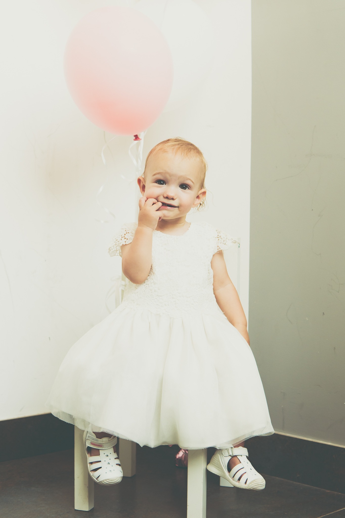 Urocza dziewczynka w białej sukience siedzi na krześle, trzyma palec w buzi, obok różowy balon.