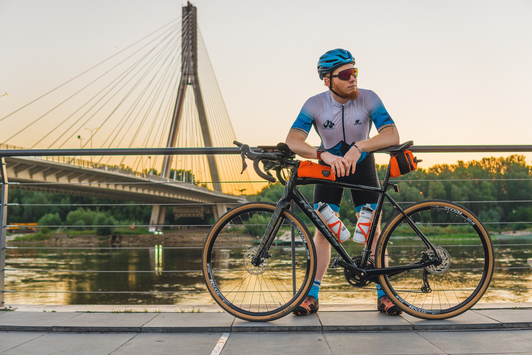 Mężczyzna w stroju rowerowym i kasku opiera się o rower na tle mostu i rzeki, w ciepłym świetle zachodzącego słońca.