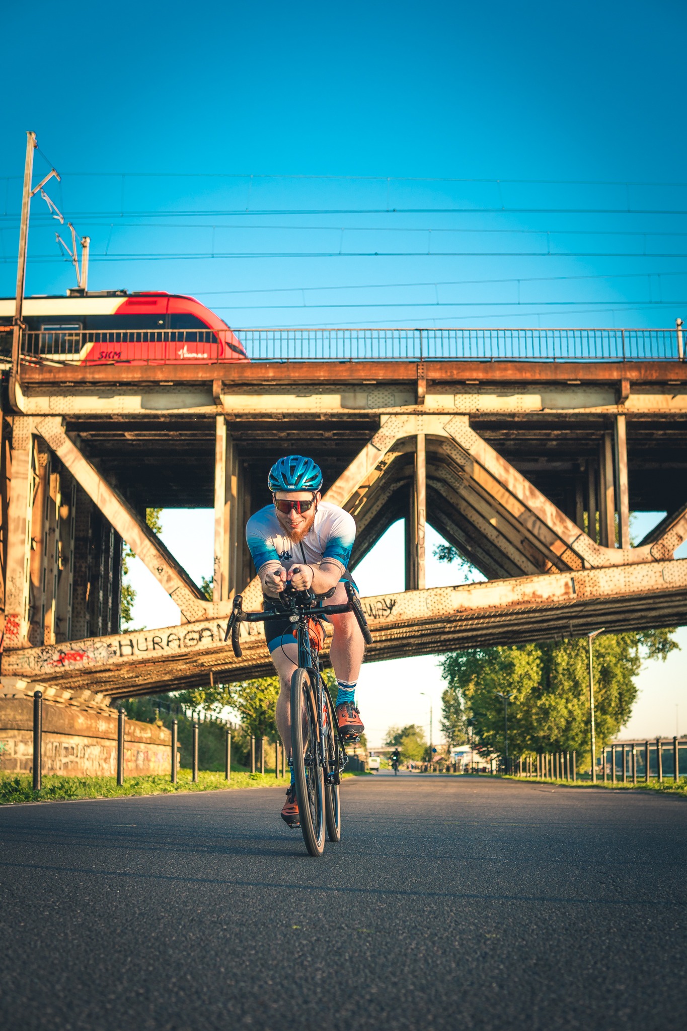Kolarz w aerodynamicznej pozycji na rowerze szosowym, pod metalowym wiaduktem z graffiti i przejeżdżającym pociągiem SKM w tle, w słoneczny dzień.