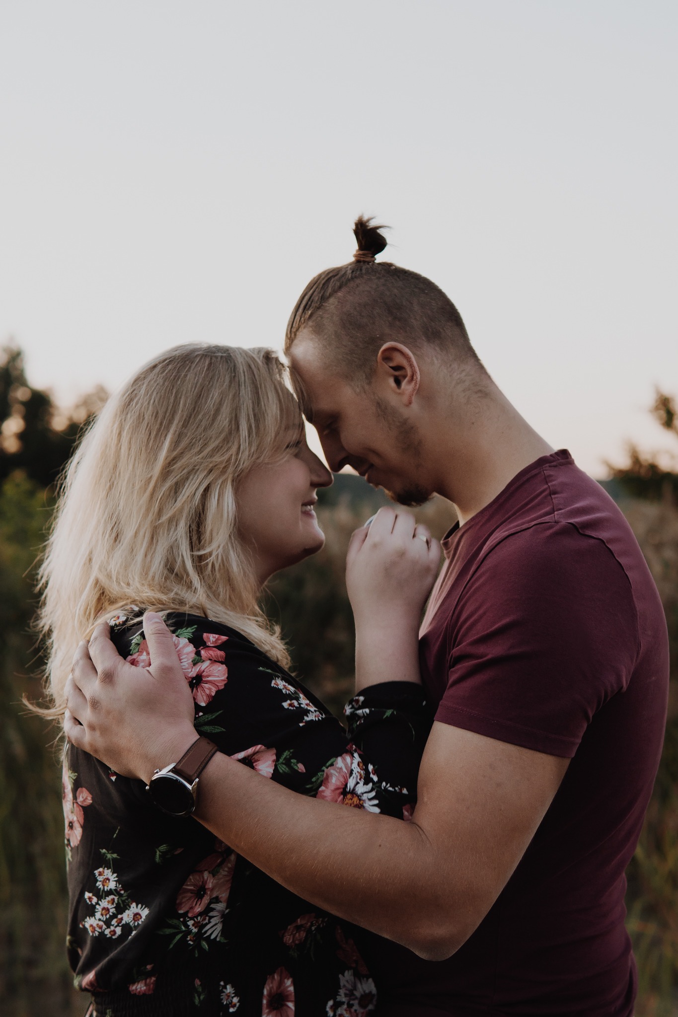 Zakochana para dotyka się czołami na tle łąki w ciepłym, naturalnym świetle; kobieta w kwiecistej sukience z zegarkiem, mężczyzna w bordowej koszulce z włosami związanymi w mały kok.
