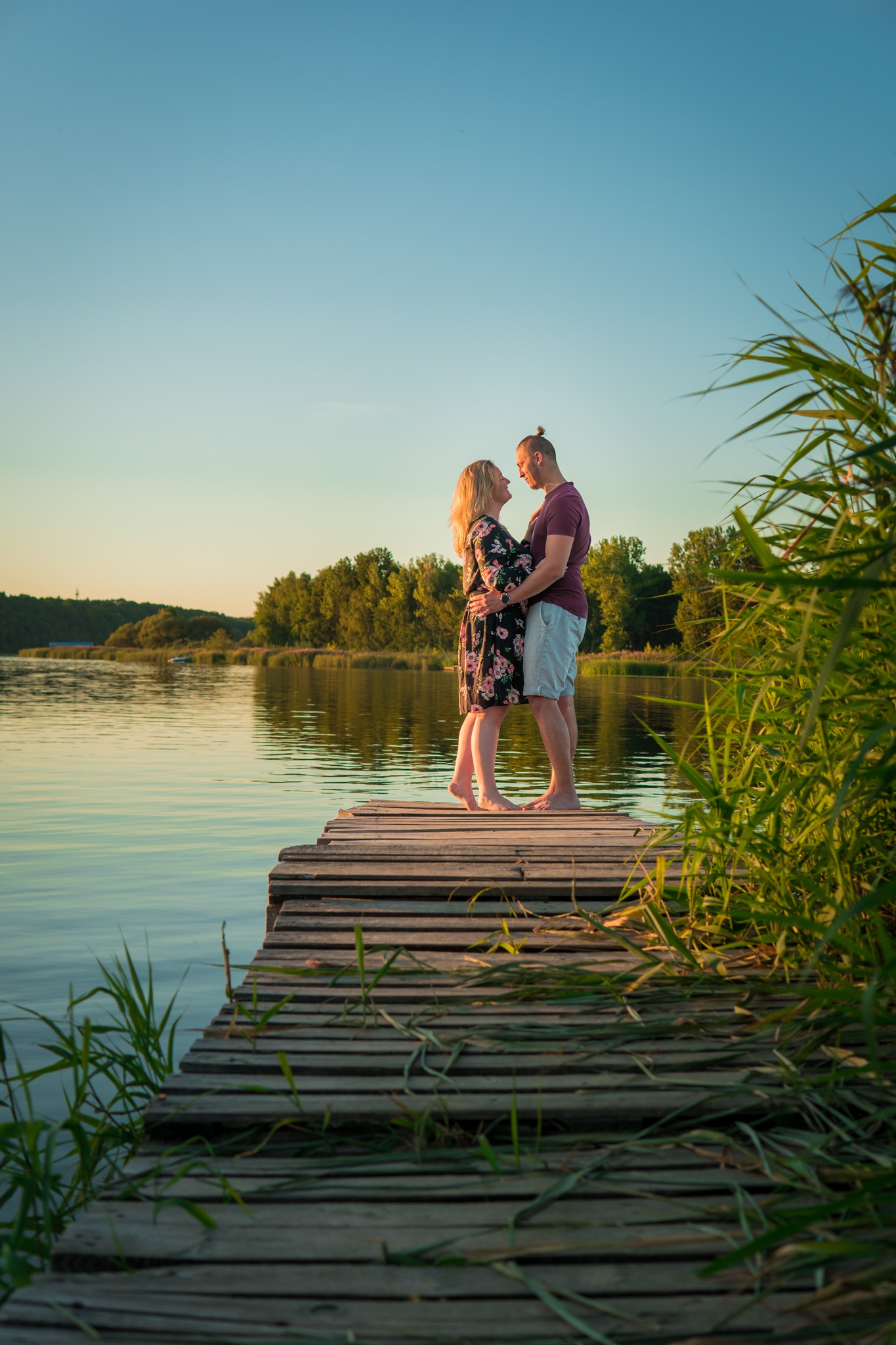 Para na drewnianym pomoście nad jeziorem o zachodzie słońca, kobieta w kwiecistej sukience obejmowana przez mężczyznę, widok na spokojną wodę i trzciny.