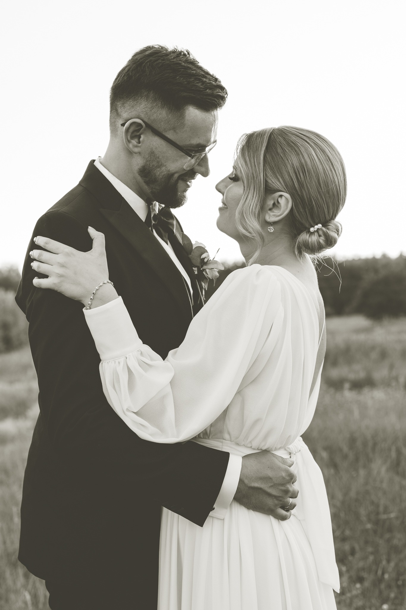 Czarno-białe zdjęcie pary młodej, obejmującej się i patrzącej sobie w oczy na tle łąki, panna młoda w sukni z bufiastymi rękawami, pan młody w garniturze i okularach.