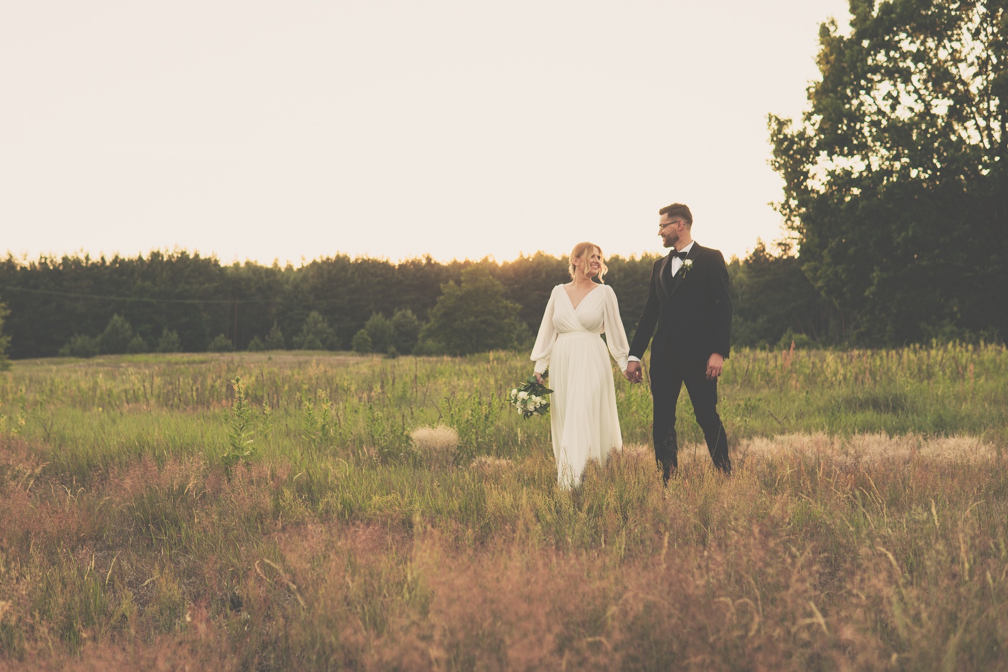 Para młoda trzymająca się za ręce spaceruje po łące w promieniach zachodzącego słońca, panna młoda w zwiewnej sukni z bukietem, pan młody w klasycznym smokingu z muszką.