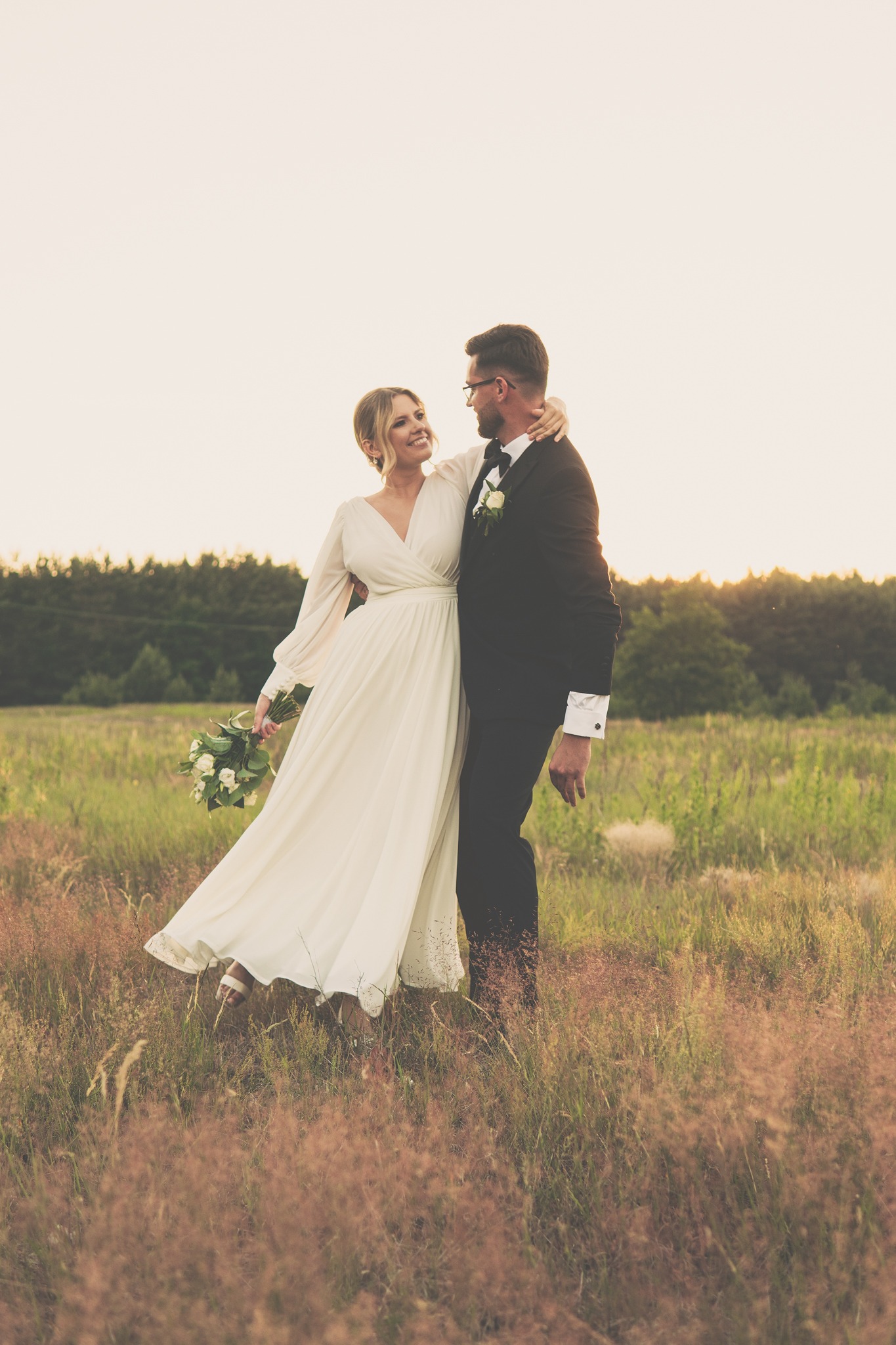 Para młoda w objęciach na łące o zachodzie słońca; panna młoda w zwiewnej sukni z bukietem białych róż, pan młody w klasycznym smokingu z muszką i butonierką.