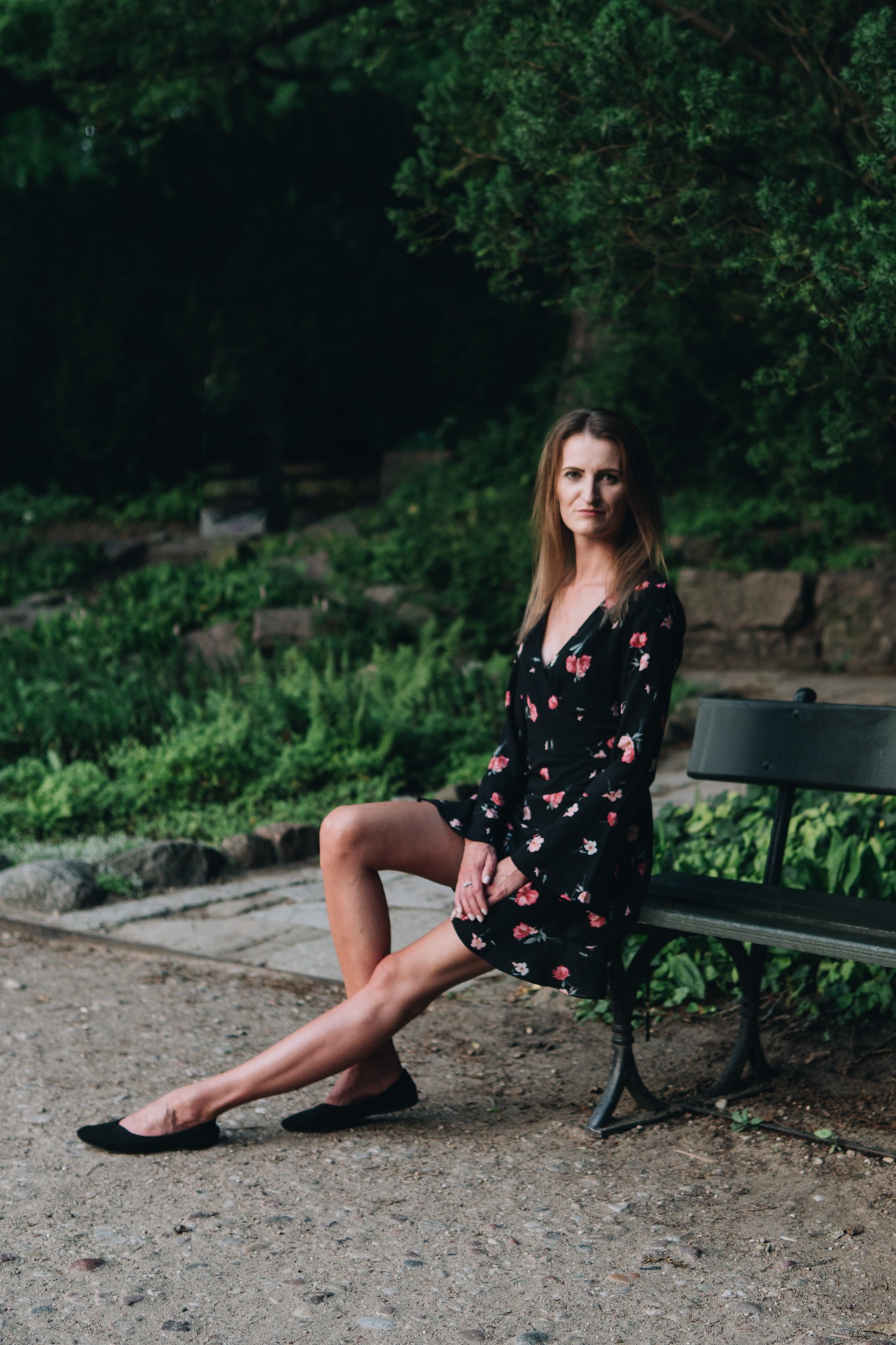 Kobieta w kwiecistej sukience siedzi na ławce w parku, z nogami skrzyżowanymi w kostkach i dłońmi splecionymi na kolanie.