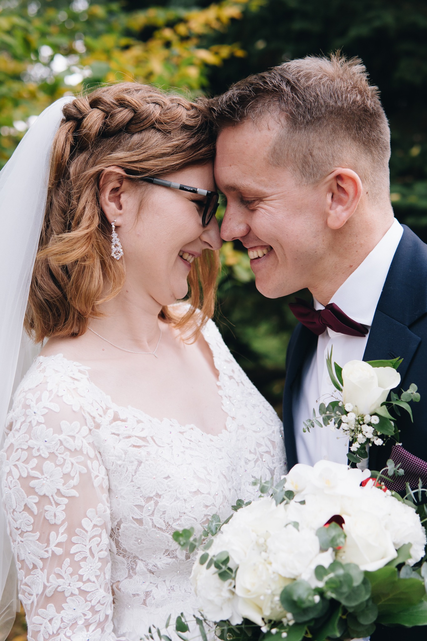 Uśmiechnięta para młoda, panna młoda w okularach, z warkoczem i welonem, pan młody w bordowej muszce, zbliżeni twarzami, bukiet z białych róż.