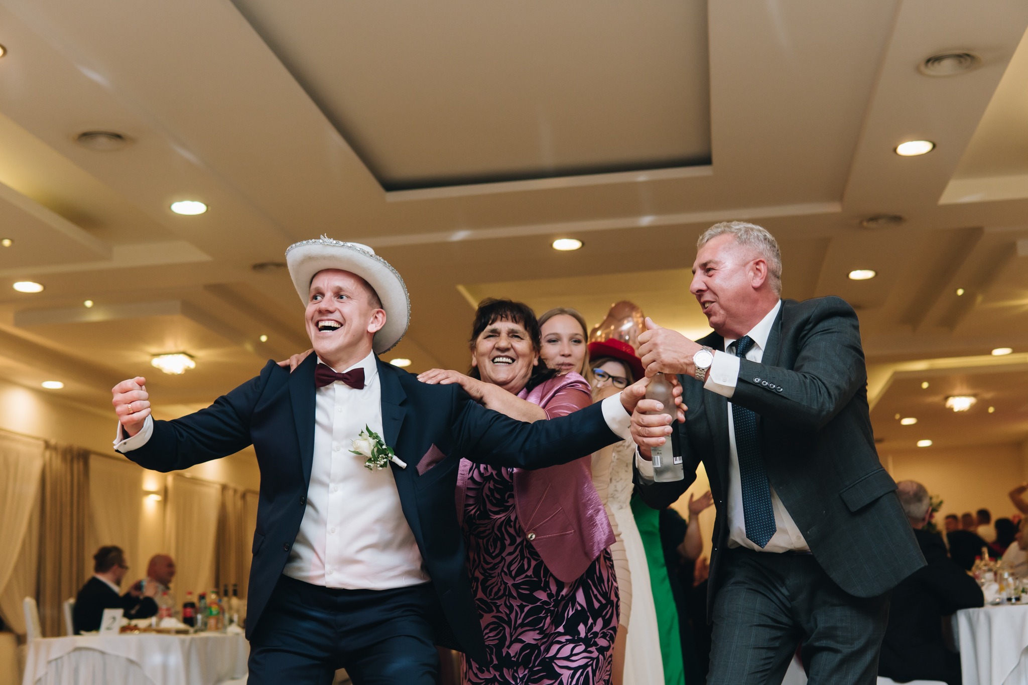 Rozbawiony pan młody w kowbojskim kapeluszu tańczy z gośćmi weselnymi, mężczyzna w garniturze otwiera butelkę wody mineralnej.