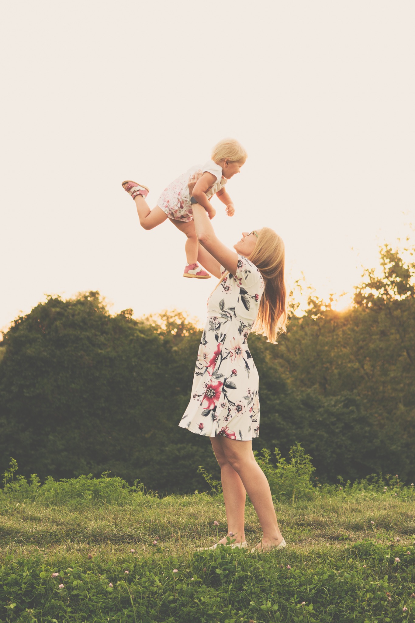 Mama w kwiecistej sukience podnosi wysoko małą blondynkę na tle zachodzącego słońca i zieleni.