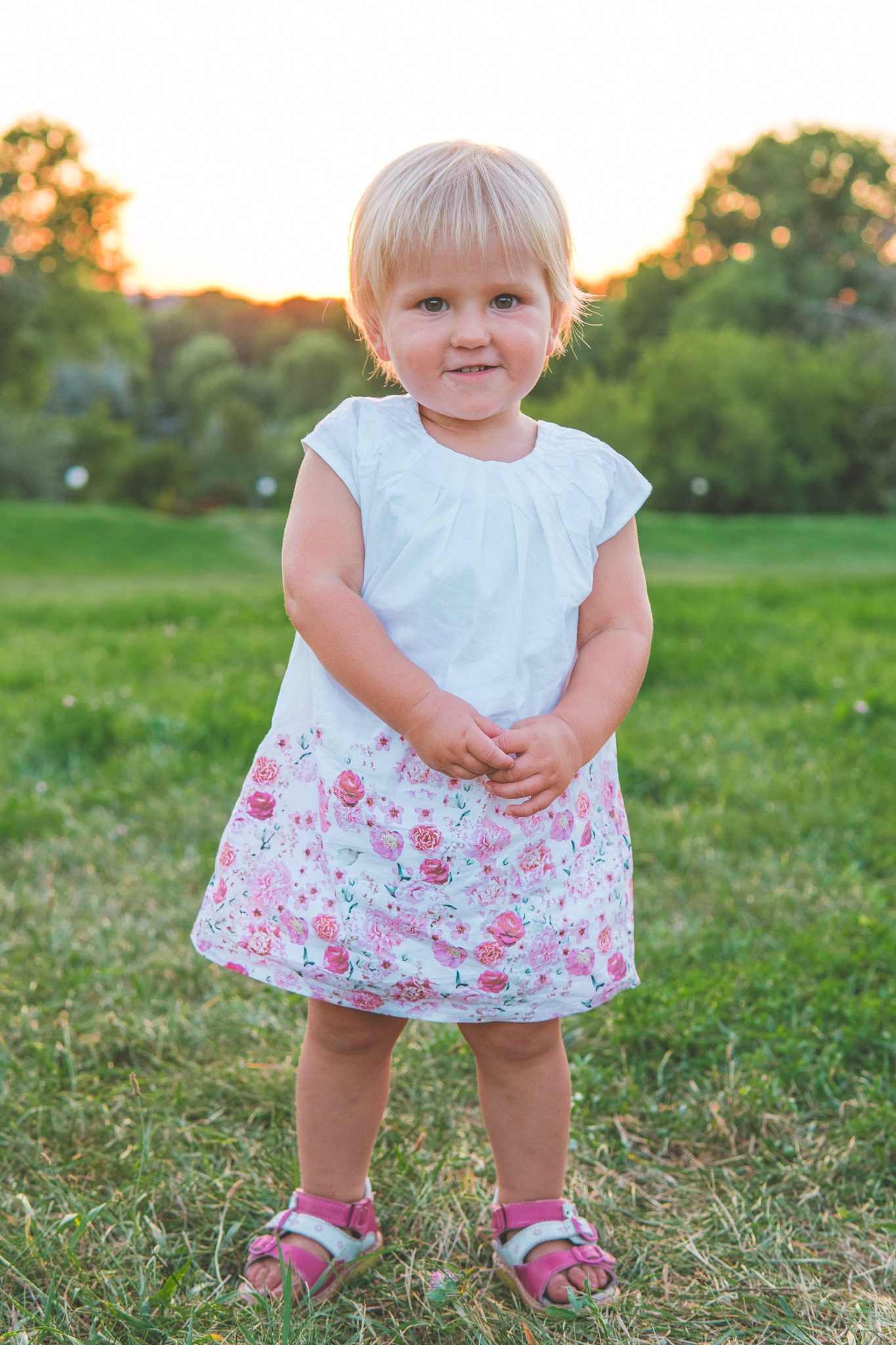 Dziewczynka w białej sukience w różowe kwiaty, stojąca na trawie, w tle drzewa i zachodzące słońce.