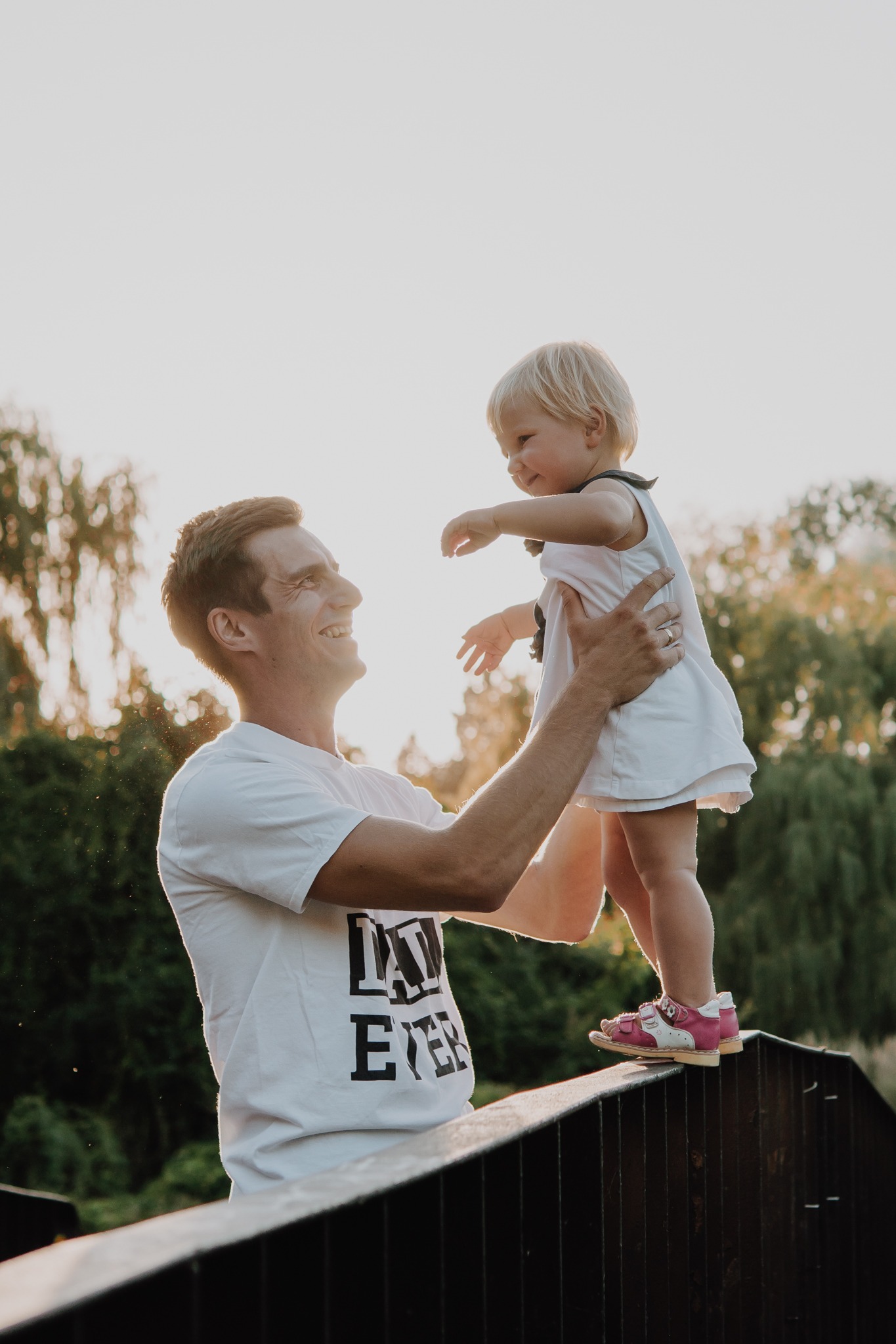 Uśmiechnięty ojciec trzymający córeczkę w białej sukience i różowych sandałach na metalowej barierce mostu, z zalesionym parkiem w tle i promieniami zachodzącego słońca.