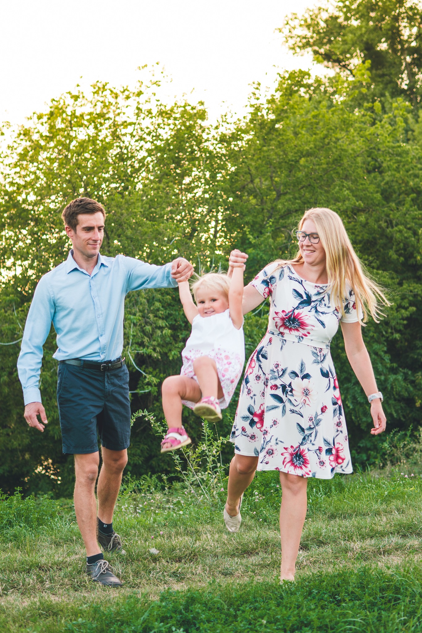 Rodzina z małą dziewczynką bawiąca się na trawie, tata i mama trzymają córkę za ręce i podnoszą ją do góry na tle zieleni.