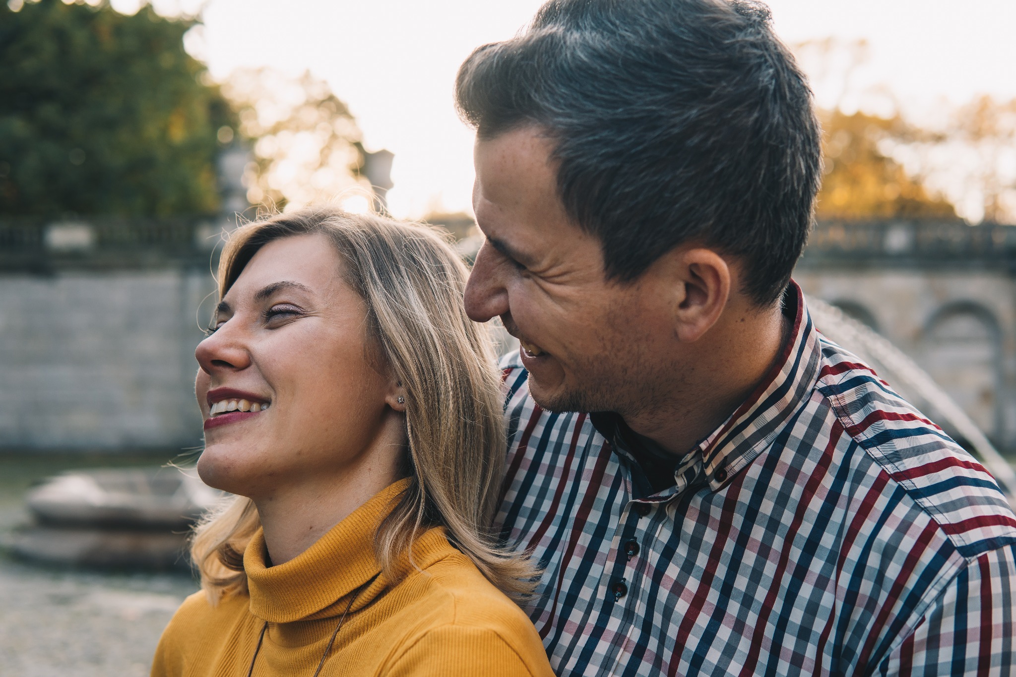 Uśmiechnięta para: blondwłosa kobieta w żółtym swetrze i mężczyzna w kraciastej koszuli, sfotografowani na tle parku z kamienną balustradą.