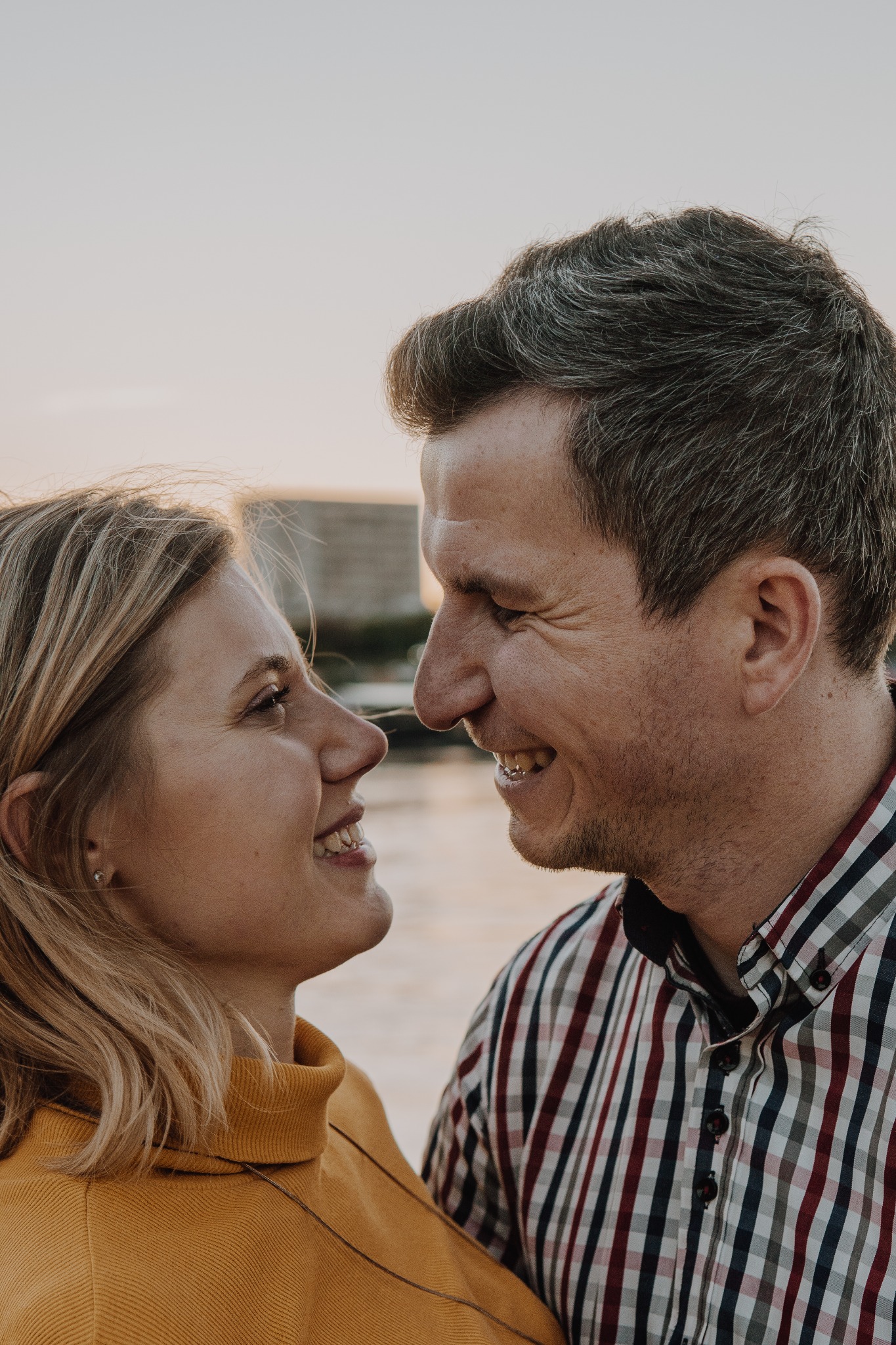 Uśmiechnięta para, kobieta w żółtym swetrze i mężczyzna w kraciastej koszuli, patrzą sobie w oczy na tle wody i betonowego nabrzeża podczas zachodu słońca.
