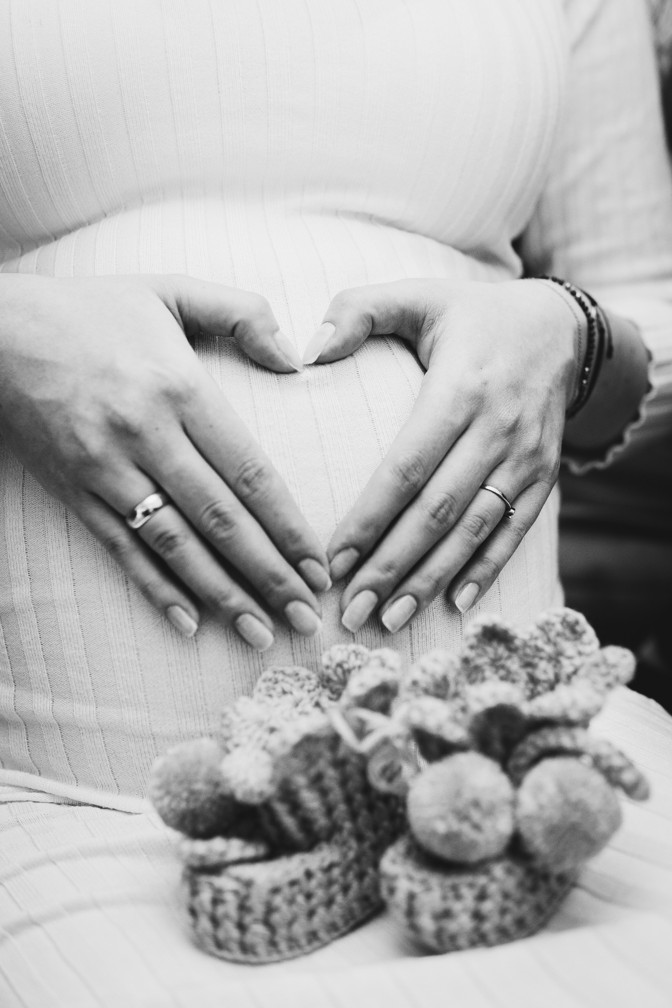 Czarno-białe zdjęcie kobiety w ciąży, dłonie ułożone w kształt serca na brzuchu, w tle dziergane buciki.