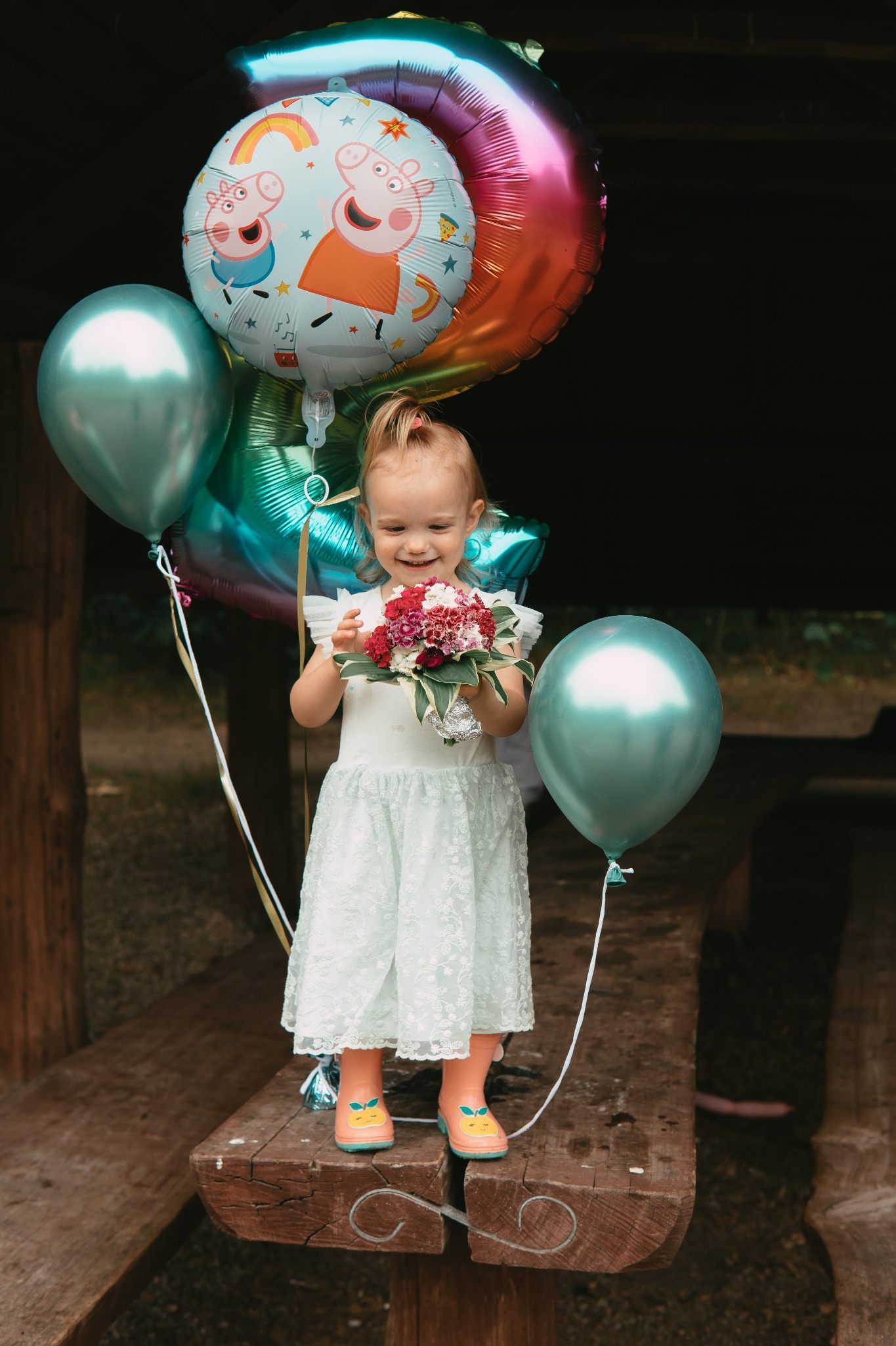 Uśmiechnięta dziewczynka w jasnej sukience trzyma bukiecik kwiatów, otoczona balonami z motywem Peppy Pig i metalicznymi balonami, stojąc na drewnianej ławie.