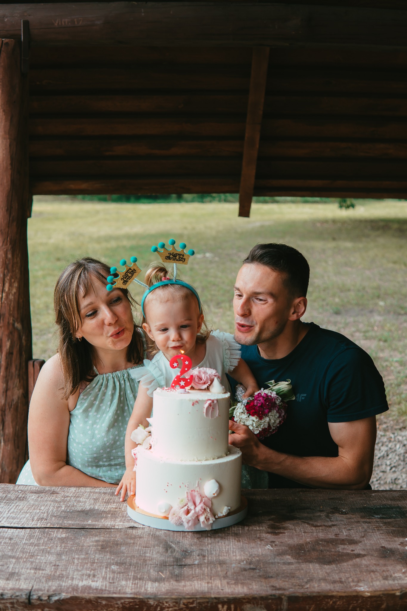 Rodzina z małą dziewczynką świętuje urodziny z dwupiętrowym tortem ozdobionym kwiatami i świeczką w kształcie cyfry dwa, na tle drewnianej altany i zieleni.