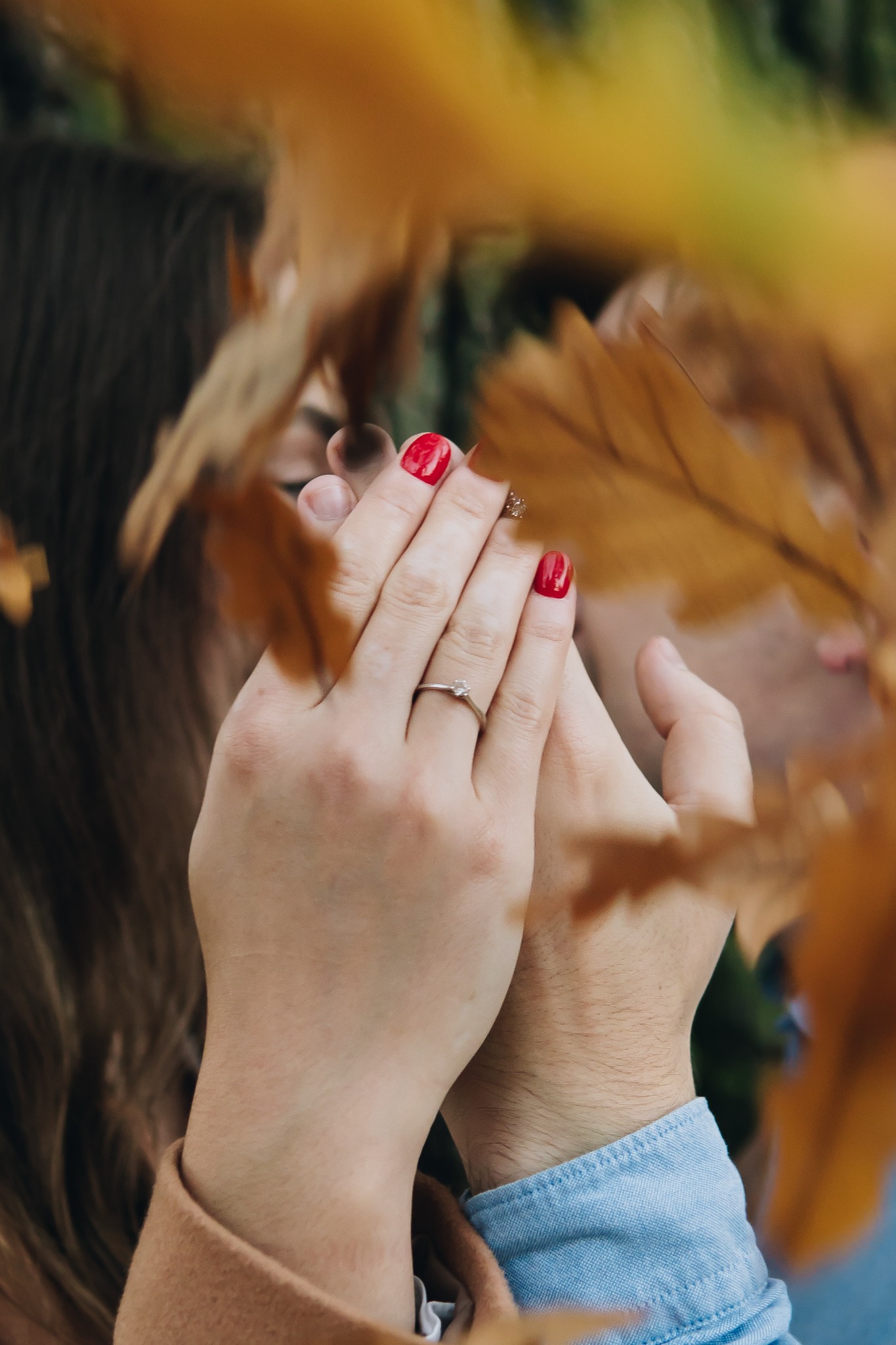 Dłonie pary z pierścionkiem zaręczynowym na tle jesiennych liści, zbliżenie na splecione palce.