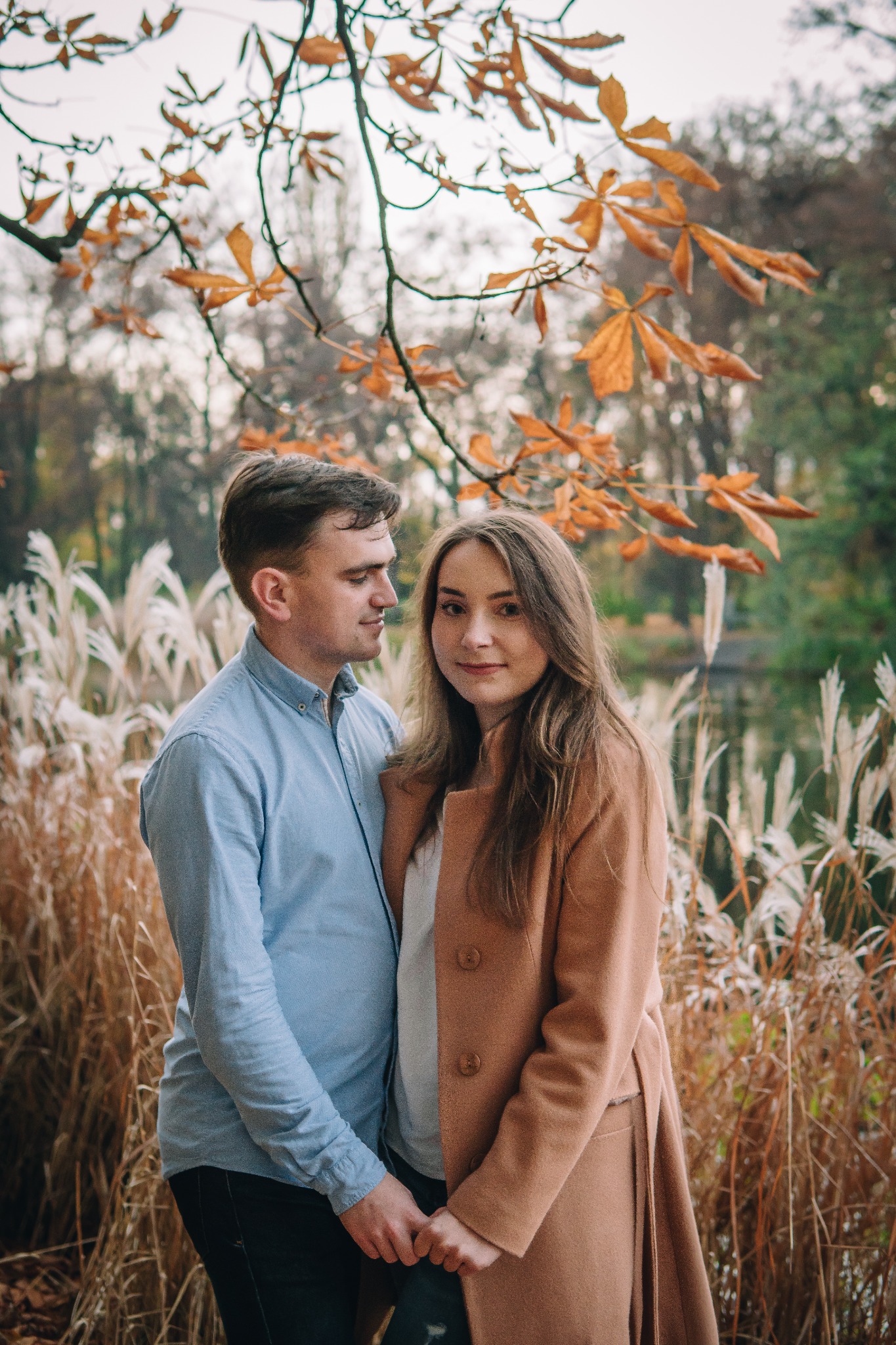 Para trzymająca się za ręce na tle jesiennego krajobrazu z trzcinami i jeziorem, gałąź z brązowymi liśćmi w górnej części kadru.