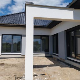 Usługi budowlane, Tomasz Wochnik, Henryk Wochnik - Nowoczesny dom jednorodzinny z szarą elewacją i dużymi oknami w czarnych ramach, widoczny fragment otynkowanej ściany z elementami wykończeniowymi przy gruncie. Teren wokół domu jeszcze...