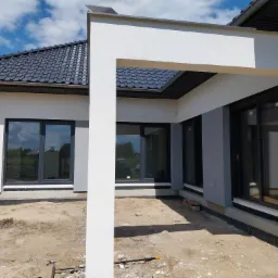 Nowoczesny dom jednorodzinny z szarą elewacją i dużymi oknami w czarnych ramach, widoczny fragment otynkowanej ściany z elementami wykończeniowymi przy gruncie. Teren wokół domu jeszcze...